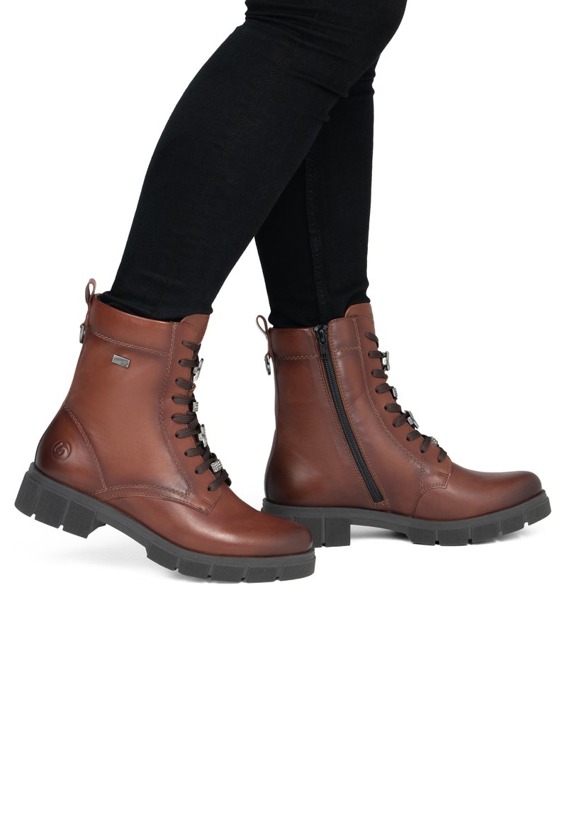 Bottines en cuir marron avec semelles noires et fermetures éclair latérales, portées avec un pantalon noir slim, vues de profil sur fond blanc.