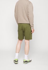 Grüne Shorts mit glatter Textur, elastischem Bund und seitlichen Taschen, kombiniert mit einem beigen Sweatshirt und hellen Sneakers.