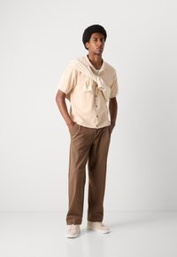 Kortærmet beige knap-op skjorte parret med løse brune bukser og cremede sneakers. En let sweater er draperet omkring skuldrene.