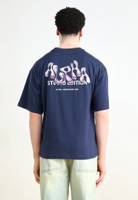 Tumši zila kokvilnas T-kreklu ar krāsainu, plūstoša stila "ALPHA" grafiku uz muguras un "STUDIO EDITION" tekstu zem tā. Īsas piedurknes.