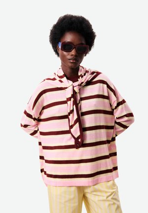 Femme portant des lunettes de soleil foncées et un haut rayé rose et marron avec une écharpe assortie, associée à un pantalon rayé jaune et blanc.