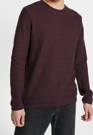 Pull bordeaux avec un motif de tricot texturé, des manches longues et un col rond. Porté par-dessus un jean noir, mettant en valeur une coupe décontractée.