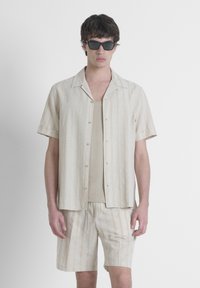 Camisa beige de manga corta con botones y rayas verticales, combinada con una camiseta sin mangas y pantalones cortos a juego, todo hecho de tela ligera.