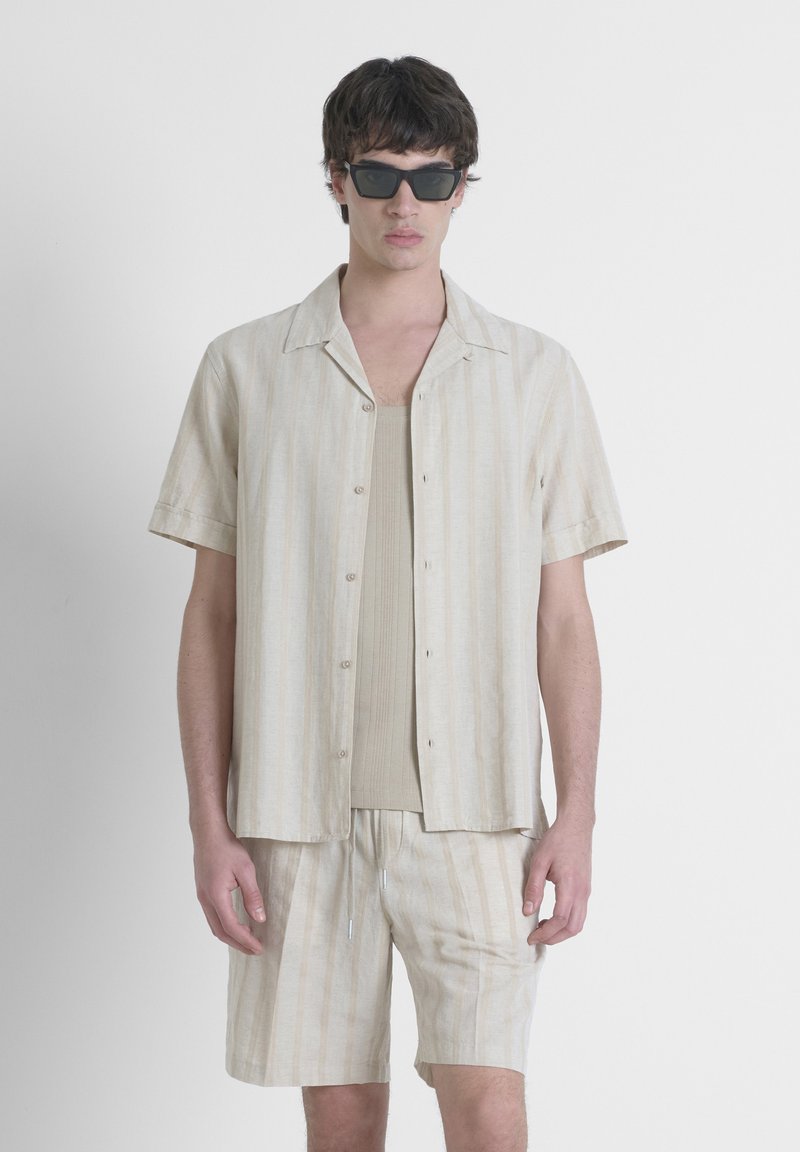 Camisa beige de manga corta con botones y rayas verticales, combinada con una camiseta sin mangas y pantalones cortos a juego, todo hecho de tela ligera.