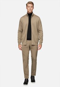 Beige knit zip-up jacket over a black turtleneck, paired with light tan pants and black sneakers, showcasing a textured design.