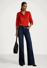 Mujer vestida con una blusa de satén roja, vaqueros de pierna ancha azul oscuro, cinturón negro, zapatos negros y sosteniendo un bolso negro.