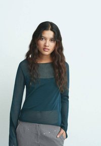 Blusa de manga larga en azul petróleo oscuro con paneles transparentes en los hombros, combinada con pantalones grises que tienen acentos plateados a lo largo de la cinturilla.