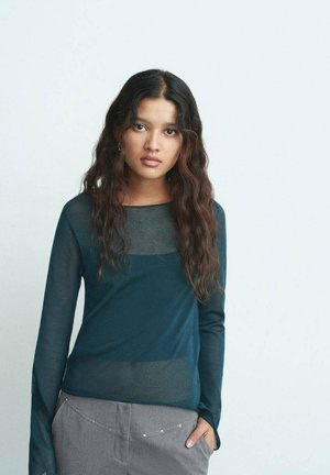 Langærmet, mørkegrøn bluse med gennemsigtige skulderpaneler, parret med grå bukser med sølvaccenter langs livremmen.