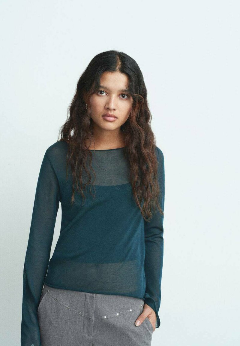 Blusa de manga larga en azul petróleo oscuro con paneles transparentes en los hombros, combinada con pantalones grises que tienen acentos plateados a lo largo de la cinturilla.