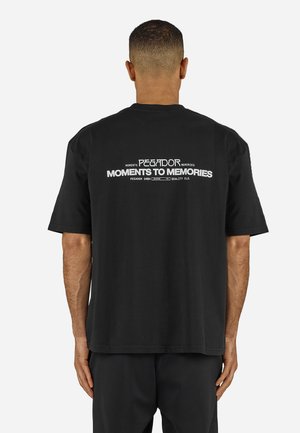 Mann iført svart T-skjorte med hvit tekst "PEGADOR MOMENTS TO MEMORIES" på ryggen, stående foran en ensfarget lys bakgrunn.