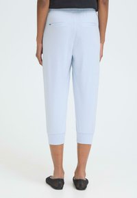 Set bagfra af en person iført lyseblå, korte bukser med elastisk talje og sorte slip-on sko, stående på hvidt gulv.