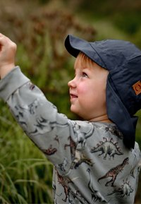 Barn som bär en grå tröja med dinosauriemönster och en mörkblå basebollkeps med öronlappar, som sträcker sig upp bland grönska.