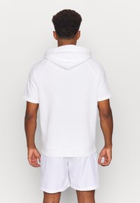 Uomo con capelli ricci corti che indossa un hoodie bianco a maniche corte e pantaloni corti bianchi, in piedi di fronte a uno sfondo chiaro e semplice.