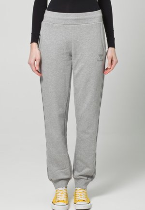 Frau trägt hellgraue Jogginghose mit Seitenstreifen, schwarzen Langarmshirt und gelbe Schnürsneaker, steht vor einem einfarbigen Hintergrund.