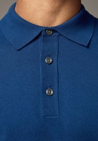 Niebieska koszulka polo z teksturowanym dzianinowym materiałem, tradycyjnym kołnierzykiem i trzema szarymi guzikami. Zbliżenie ukazujące szczegóły przeszyć.
