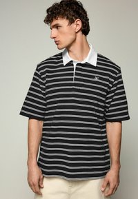 Stribet polo shirt i sort og hvid med hvid krave, korte ærmer og et lille broderet logo på brystet. Lavet af bomuldsmateriale, afslappet pasform.