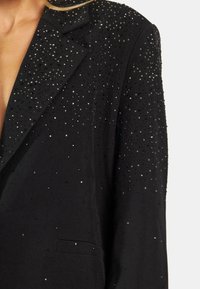 Blazer negro con un diseño ajustado, adornado con rhinestones dispersos, que presenta un cuello muesca y un único bolsillo discreto.