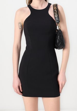 Woman wearing a sleeveless, fitted black dress with white trim around the neckline, holding a black quilted handbag with gold details.