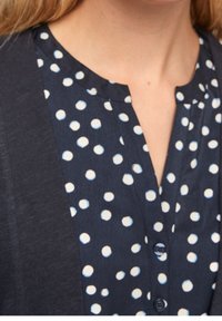 Marinblus med vita prickar, design med knapprad fram, låg halsringning och kontrasterande tygkant på sidorna. Mjuk textur.