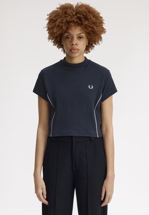 Femme aux cheveux bouclés portant un t-shirt court bleu marine avec un liseré blanc et un pantalon plissé noir, debout devant un fond uni.