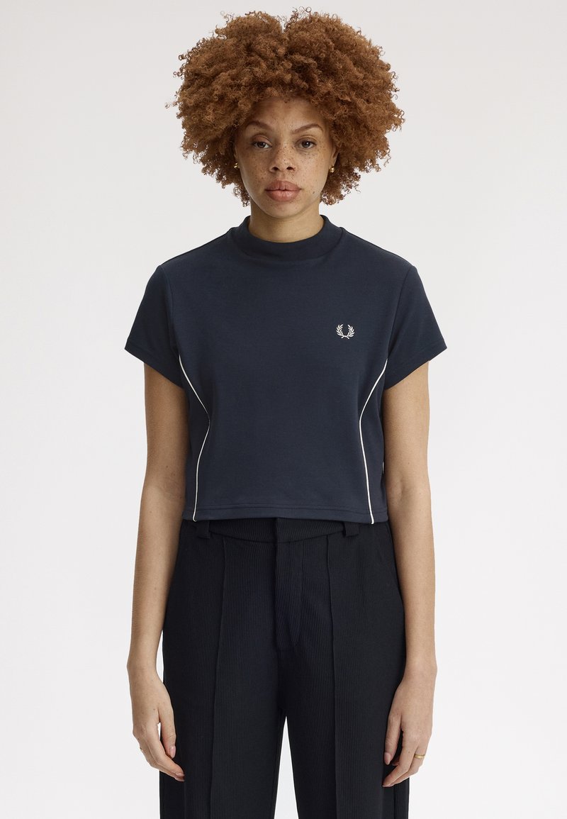 Femme aux cheveux bouclés portant un t-shirt court bleu marine avec un liseré blanc et un pantalon plissé noir, debout devant un fond uni.