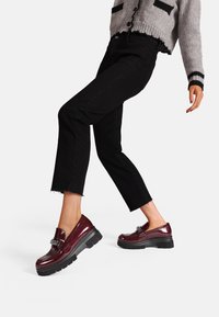 Mocassini in pelle verniciata bordeaux con suola in gomma nera spessa e un accento decorativo a forma di fibbia. Indossati con jeans neri a vita bassa.