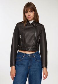 Kobieta nosząca krótka czarna skórzana kurtka z detalami w postaci zamka i paska, w zestawieniu z dżinsami o wysokim stanie w odcieniu niebieskim, na jednolitym tle.