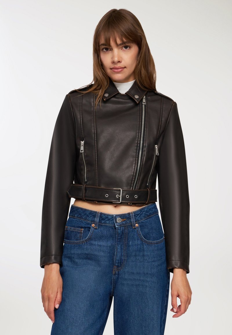 Kobieta nosząca krótka czarna skórzana kurtka z detalami w postaci zamka i paska, w zestawieniu z dżinsami o wysokim stanie w odcieniu niebieskim, na jednolitym tle.