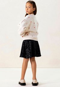 Lichtgrijze sweatshirt met kleurrijke borduursels van harten en sterren, gecombineerd met een zwarte paillettenrok en beige schoenen met zwarte zolen.