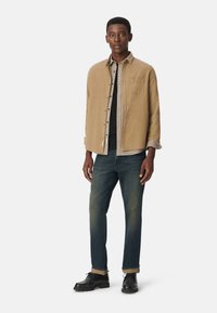 Jasnobrązowa koszula zapinana na guziki nałożona na koszulę w kratę, połączona z ciemnymi dżinsami i czarnymi butami. Posiada podwinięte rękawy i kieszeń na piersi.