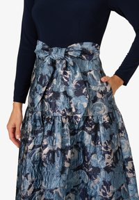 Rock mit Blumenmuster in Blau- und Metallictönen, mit einer großen Schleife an der Taille und seitlichen Taschen. Strukturierter Stoff.