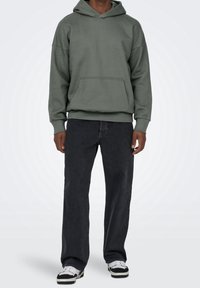 Sweat à capuche vert avec poche avant, coupe décontractée et capuche à cordon réglable, associé à un jean noir droit et à des baskets noir et blanc.