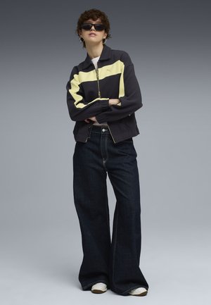 Person i sort og gul lynlåsjakke, vide mørke denimjeans, hvide sneakers, solbriller og øreringe med hoops, der står foran en grå baggrund.