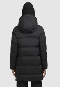 Jachetă puf cu glugă, de culoare neagră, având secțiuni matlasate și un design de lungime medie. Acoperită cu o căciulă tricotată neagră.