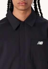 Fekete gombos ing, Strukturált gallérral, mellzsebből és fehér New Balance logóból a bal felső oldalon. Simább textúrával.