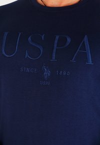 Navy modra majica iz mehkega materiala z dvignjenim napisom "USPA" in majhnim logotipom, okroglim izrezom in kratkimi rokavi.
