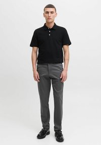 Polo noir à manches courtes avec trois boutons, associé à un pantalon gris ajusté. Le mannequin porte des chaussures noires, se tenant devant un fond uni.