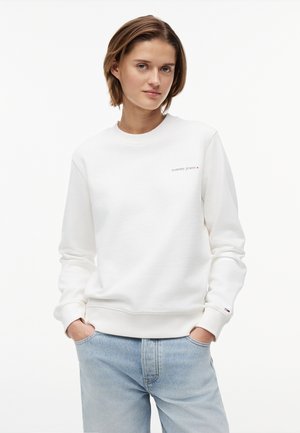 Mladá osoba s krátkymi hnedými vlasmi, oblečená v bielej mikine Tommy Jeans a svetlomodrých džínsoch, ruky vo vreckách, neutrálne pozadie.