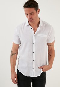 Camisa blanca de manga corta en tejido texturizado con botones negros, un bolsillo en el pecho y puños doblados, combinada con jeans oscuros.