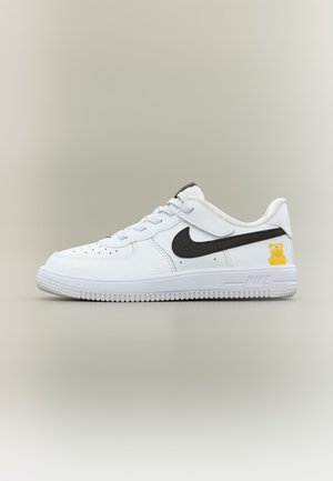 Weißer Sneaker mit niedrigem Schaft, schwarzem Nike-Swoosh, Klettverschluss und kleinem gelbem Bärenmotiv nahe der Ferse auf neutralem Hintergrund.
