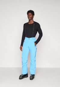 Šviesiai mėlynos sniego kelnės su petnešomis ir užtraukiamomis kišenėmis, dėvimos virš juodos ilgomis rankovėmis marškinėlių. Stilius pritaikytas žiemos lauko veiklai.