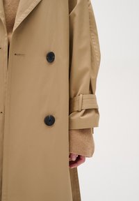 Beige trench coat featuring large black buttons, a belted sleeve detail, and smooth texture, worn over a beige sweater.