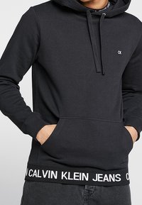Muž v černé mikině Calvin Klein s přední kapsou a logem na hrudi a pase, s rukama v kapsách.