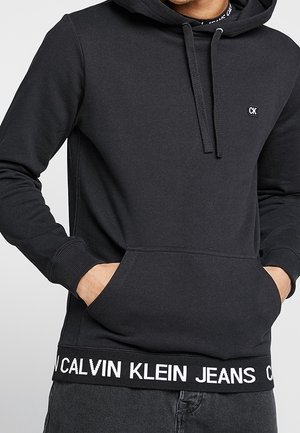 Uomo che indossa una felpa nera Calvin Klein con tasca frontale e logo sul petto e sull'orlo, mani in tasca.