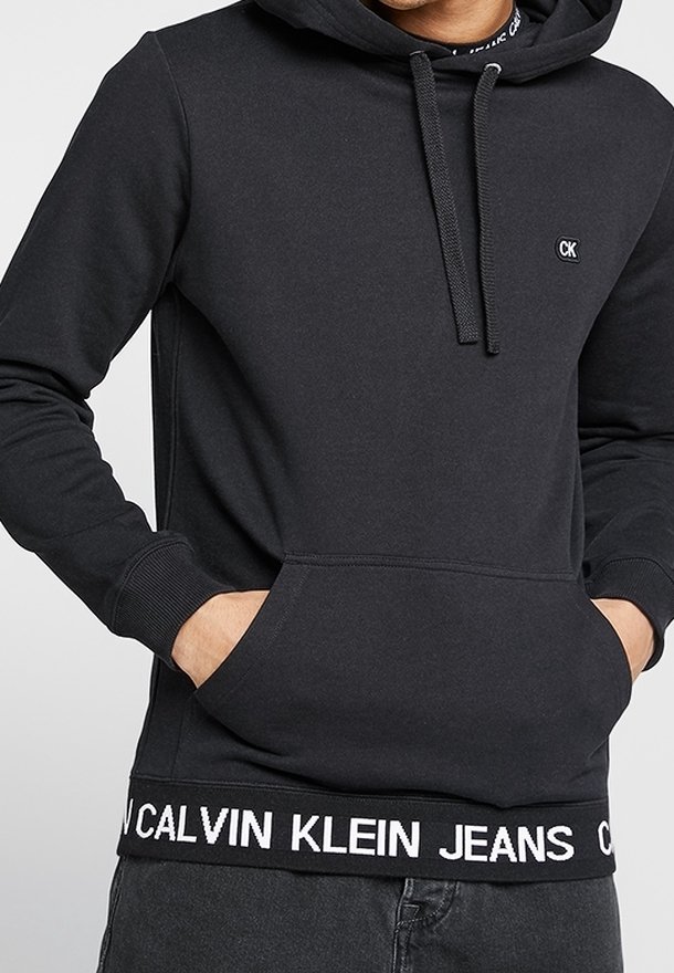 Muž v černé mikině Calvin Klein s přední kapsou a logem na hrudi a pase, s rukama v kapsách.