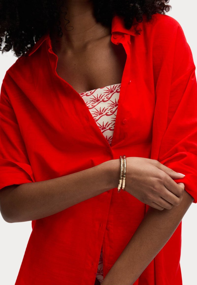 Camicia rossa con bottoni e maniche corte sopra un top a fantasia, impreziosita da braccialetti sovrapposti oro e beige su un braccio di carnagione scura.