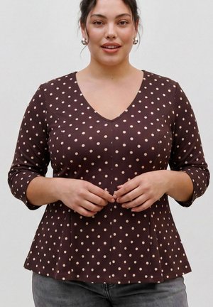 Femme portant une chemise marron à manches longues avec de légers pois clairs et des boucles d'oreilles rondes en argent, debout devant un fond uni clair.