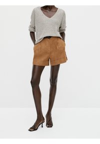 Un pull en maille gris clair à manches roulées, associé à un short en lin marron avec des plis et des poches latérales. Des sandales à talons strappées noires.