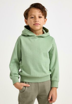 Felpa verde chiaro con cappuccio, tasca a marsupio e polsini a coste. Tessuto morbido con vestibilità rilassata, abbinata a pantaloni beige.