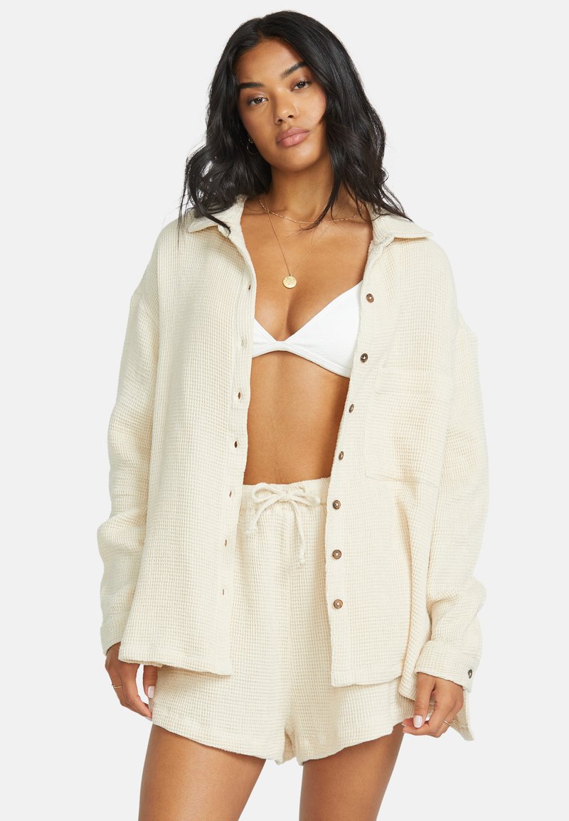Camicetta in crema a trama waffle con bottoni e taschino davanti, abbinata a pantaloni corti con coulisse coordinati. Vestibilità casual e rilassata.
